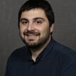 Stelios's headshot; brown hair, brown eyes, brown stubble, navy blue button down